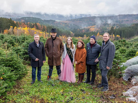 Gruppenfoto im Christbaumdorf Spessart bei Christbaum-Klug Sieben Personen, darunter die Christbaumkönigin im rosa Kleid, stehen in einer Weihnachtsbaumplantage im Spessart