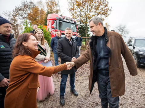 Begrüßung von Markus Söder im Christbaumdorf Mittelsinn Markus Söder schüttelt einer Frau vor mehreren Menschen die Hand auf einem Hof mit Fahrzeugen im Hintergrund