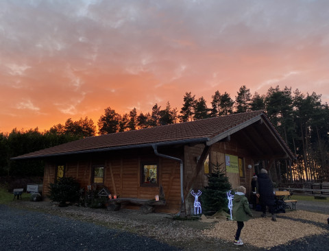 Holzhütte im Christbaumdorf Klug, Treffpunkt für Firmenweihnachtsfeier mit Christbaum selbst sägen Holzhütte bei Sonnenuntergang vor Nadelbäumen, Menschen stehen davor und bereiten eine Firmenweihnachtsfeier vor bei der Christbäume selbst gesägt werden