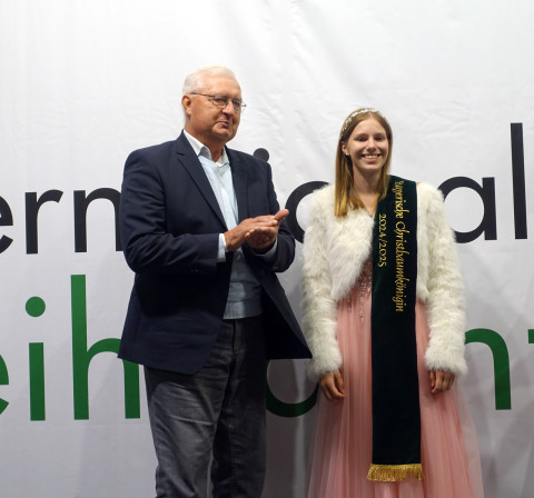 Bayerische Christbaumkönigin Sina Klug 2024 / 2025 wird geehrt Eine junge Frau mit Krone und Schärpe steht in einem rosa Kleid neben einem älteren Mann auf einer Bühne